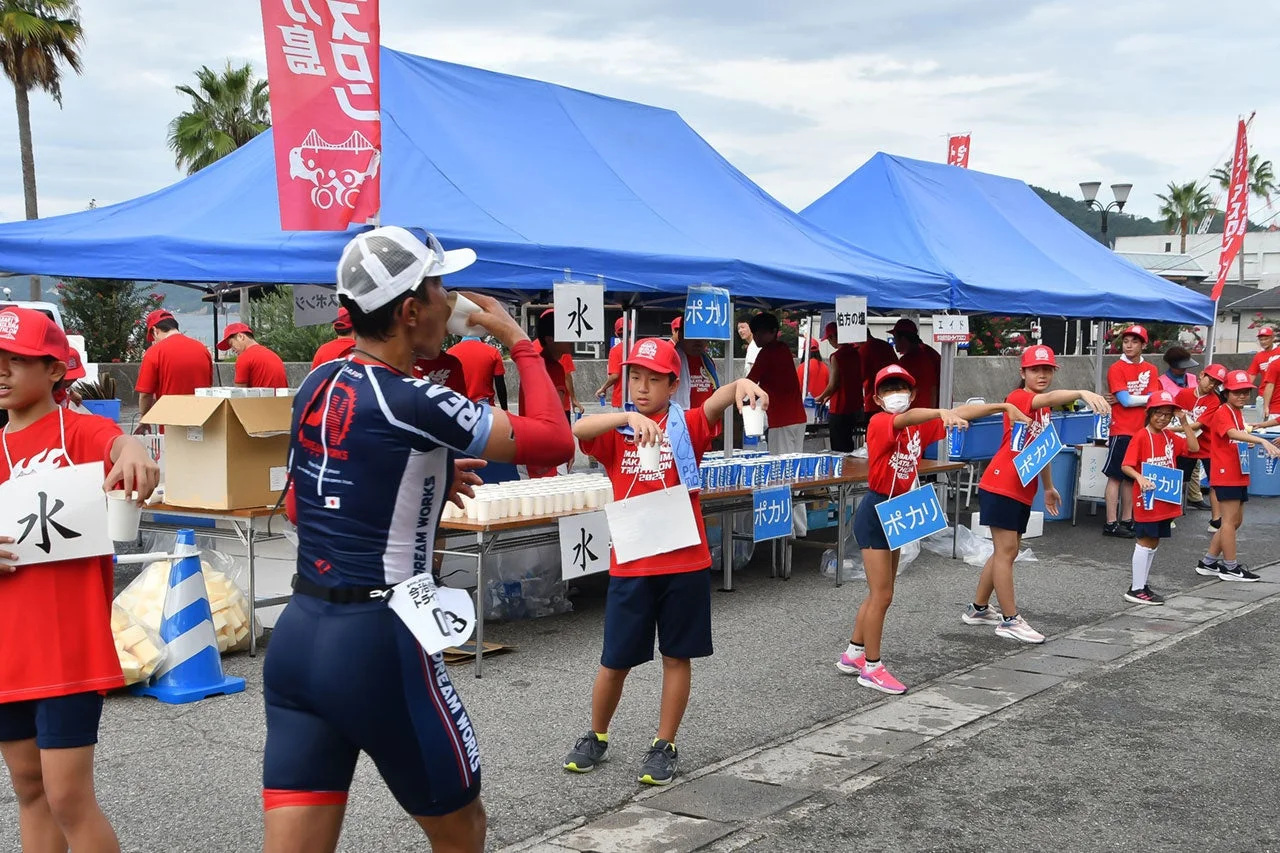 トライアスロン大会のエイドステーションで水分補給をする参加者とボランティアの子供たち