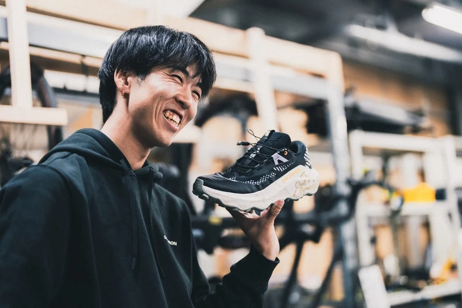 黒いフーディーを着た男性が笑顔で黒と白のスポーツシューズを手に持っています。