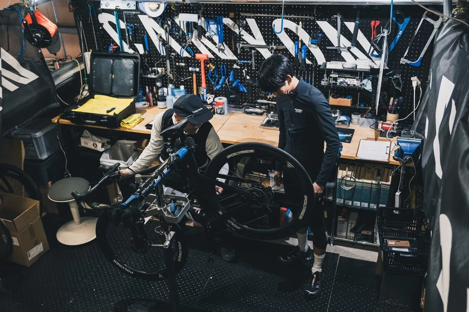 自転車の整備工場で、二人の男性がロードバイクのメンテナンスをしている様子。