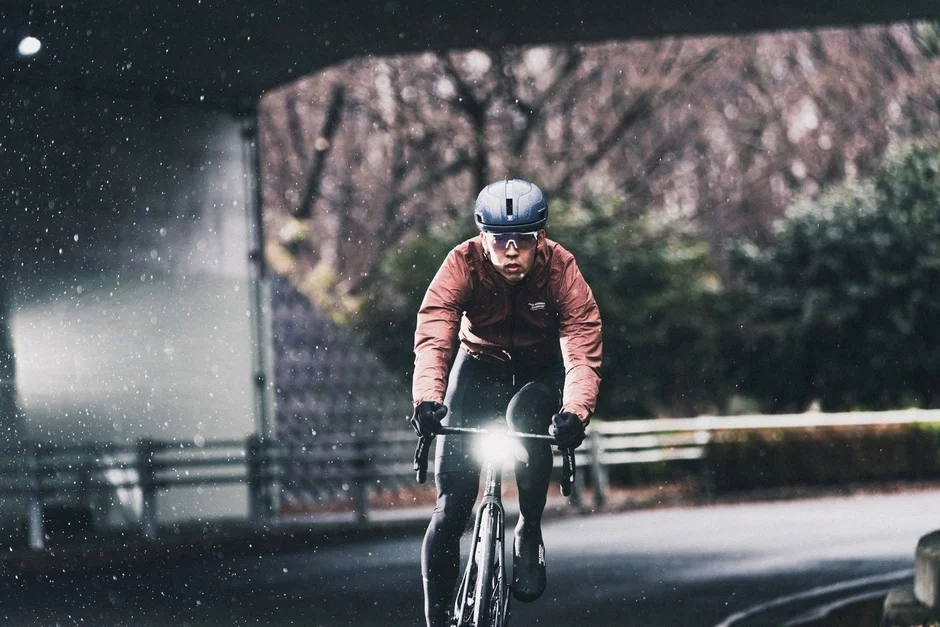 ヘルメットとサングラスを着用したサイクリストが、雨または雪が降る中でロードバイクに乗っています。