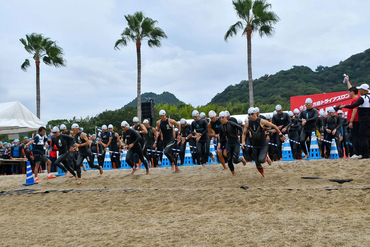 砂浜でトライアスロン競技がスタートし、ウェットスーツを着た多くの選手が一斉に走り出す様子