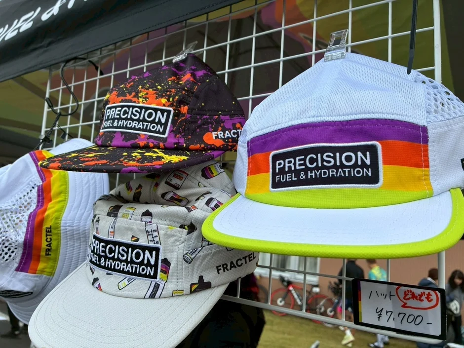ワイヤーラックに「PRECISION FUEL & HYDRATION」と「FRACTEL」のロゴが入ったカラフルなキャップが複数展示されている。価格は一律7,700円と表示されており、屋外での販売イベントの様子を捉えた画像である。