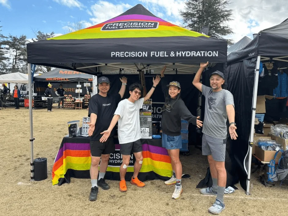 Precision Fuel & Hydrationのテント前で男女4人が笑顔でポーズ。アウトドアイベントのブースで、スポーツ関連の展示会と思われる活気ある雰囲気。