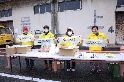 黄色のジャンパーを着た4人の女性が屋外で「いっぺーまーさん」と書かれた横断幕を持って立っています