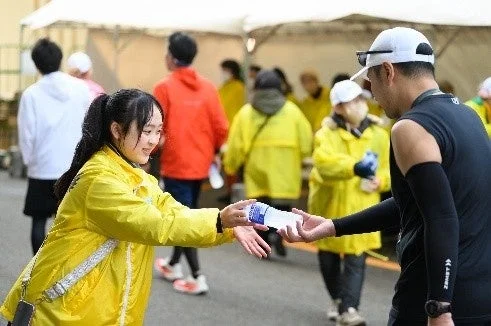 スポーツイベントの給水所で、黄色いジャケットを着たボランティアの女性が、ランナーにペットボトルの水を笑顔で手渡している場面