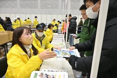 屋内会場で、黄色いジャケットを着たスタッフが、マスクを着用した参加者に対して資料を配布している場面