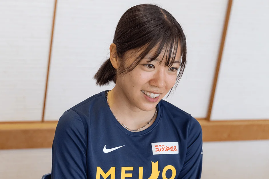 紺色のスポーツウェアを着た若い女性が、穏やかな笑顔で座っています。室内でリラックスした雰囲気の写真です。