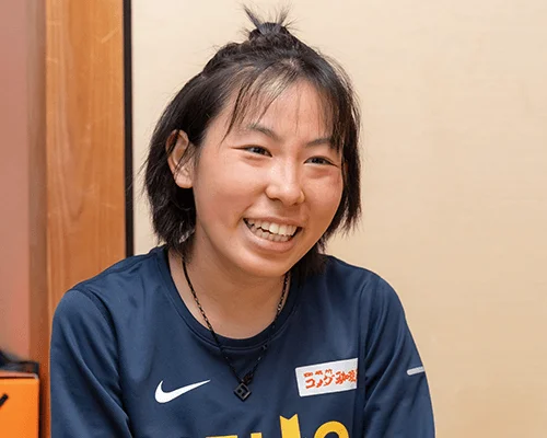 若々しい日本人女性が満面の笑みを浮かべています。髪はちょんまげのように結ばれ、紺色のスポーツウェアにはナイキのロゴとコカ・コーラの文字が見えます。