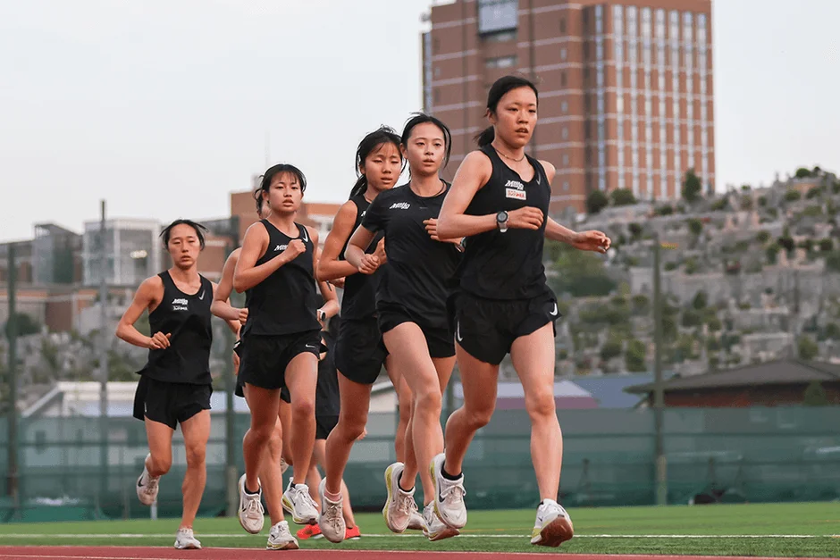 陸上競技場で黒いウェアを着用した複数の女性ランナーたちが、真剣な表情で走っています。チームでのトレーニング風景と見られます。