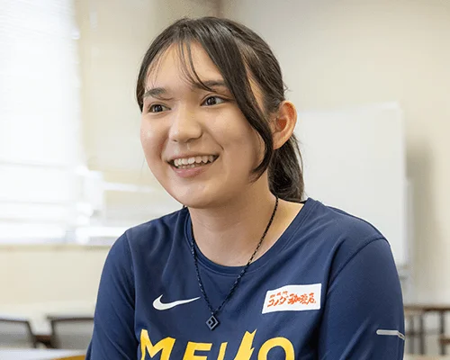 紺色のスポーツウェアを着た若い女性が笑顔で座っている。シャツには「MEIJO」と「ランニング専門店」の文字が見える。明るい室内で、楽しそうな表情が印象的。
