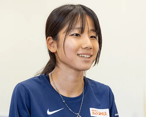 紺色の長袖シャツを着た若いアジア人女性のポートレート。彼女は笑顔を見せており、髪は後ろでまとめられています。シャツにはナイキのロゴと「コジマ薬局」の文字が見えます。