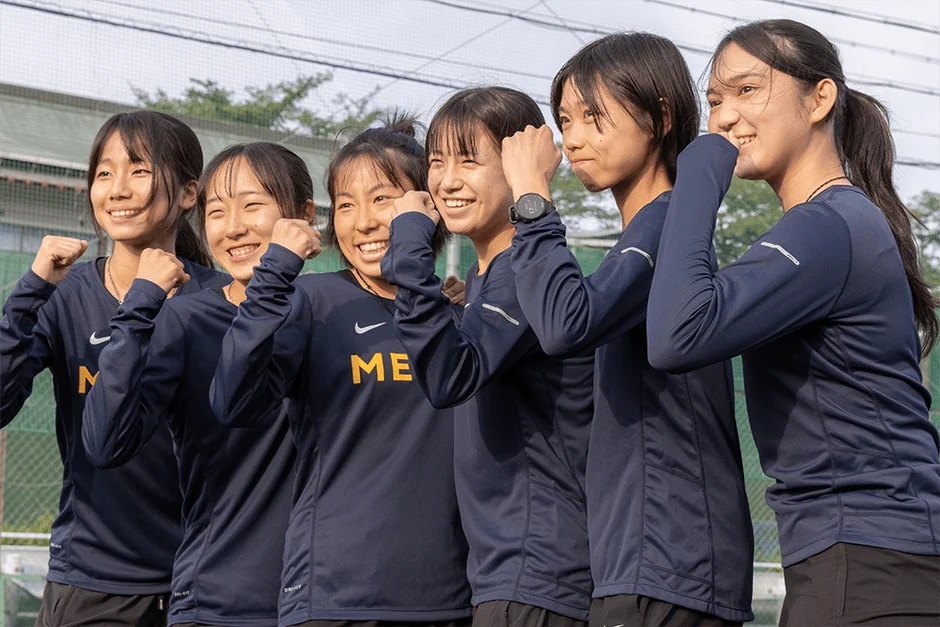 紺色の長袖スポーツウェアを着た5人の若い女性たちが、笑顔で拳を握り、ガッツポーズをしている写真です。チームの一体感や勝利への意気込みが感じられ、明るく活動的な雰囲気が伝わってきます。