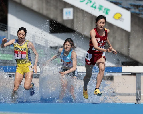 陸上競技の障害物競走で、3人の女子選手が水濠を越える瞬間を捉えた写真です。水しぶきを上げながら力強く走る姿から、競技の激しさと躍動感が伝わってきます。