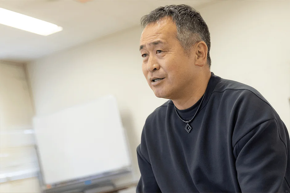 グレーがかった髪の中年アジア人男性が、黒いカジュアルなシャツとネックレスを着用し、屋内で会話している様子です。真剣な表情で右方向を見ており、背景には白いボードが見えます。