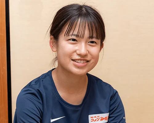 笑顔の若い女性がカメラ目線で写っています。暗い色の半袖シャツを着用し、背景はシンプルな壁です。室内で撮影されたポートレート写真です。