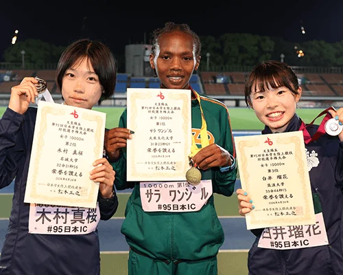 「天皇賜杯第95回日本学生陸上競技対校選手権大会」女子10000mの表彰式で、1位サラ・ワンジル選手、2位木村真桜選手、3位白井瑠花選手がメダルと賞状を手に笑顔で写っています。