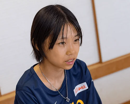 青いスポーツウェアを着たショートヘアの若い女性が、真剣な表情で視線を送っている。シャツには「がんばろう、熊本。」の文字があり、背景は和室のような雰囲気である。