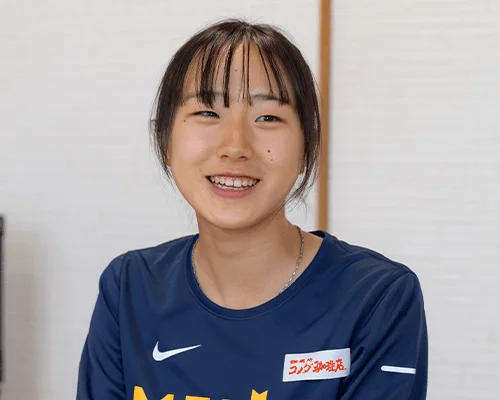 笑顔でカメラを見つめる若いアジア人女性のポートレートです。彼女は紺色の長袖シャツを着用しており、胸元には「ラングドシャ」の文字とナイキのロゴが見えます。