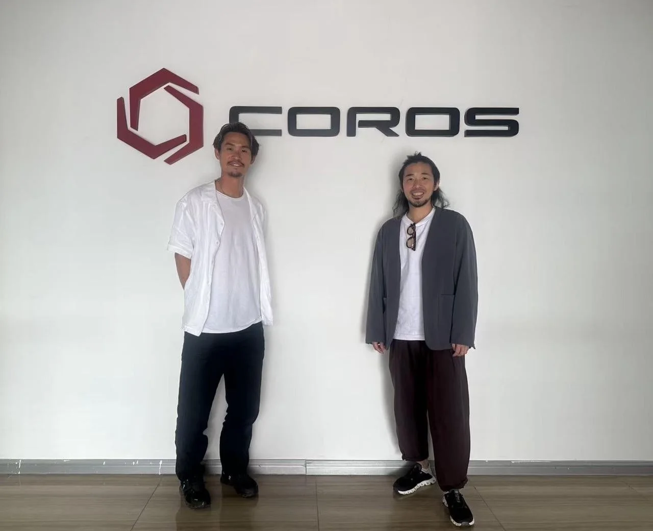 白壁の「COROS」ロゴの前で、カジュアルな服装の男性2人が笑顔で立っている写真です。