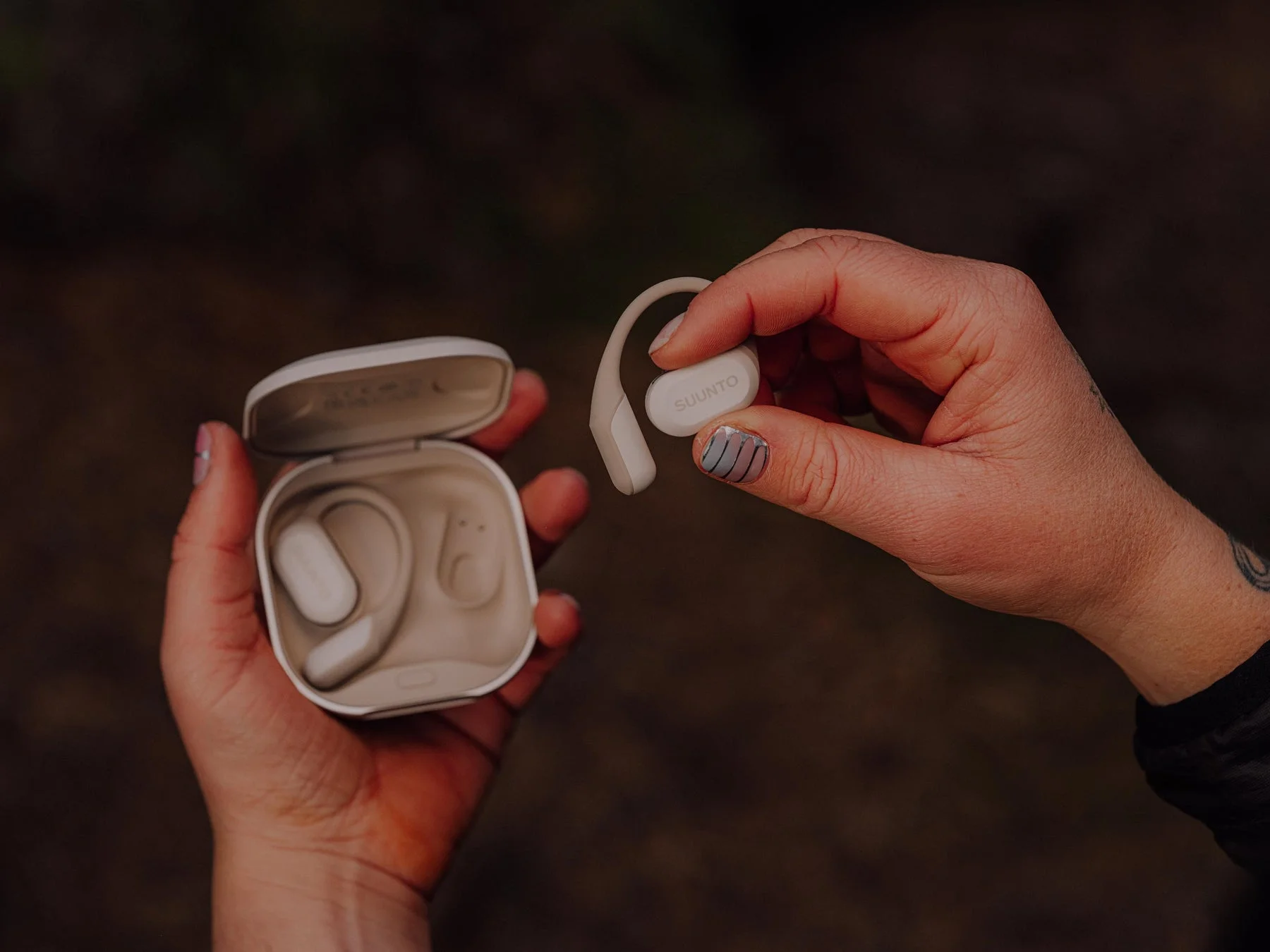 女性がSuuntoのワイヤレスイヤホンと充電ケースを両手に持っています。耳掛け式のイヤホンはベージュトーンで、シンプルかつスタイリッシュなデザインが特徴です。