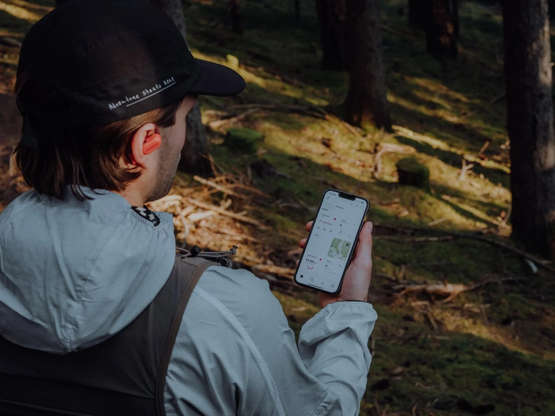 森の中でキャップをかぶった人物がスマートフォンを見ている。耳にはイヤホンを装着し、スマホ画面にはフィットネスアプリが表示されている。アウトドアでの活動と健康管理を示す一枚。